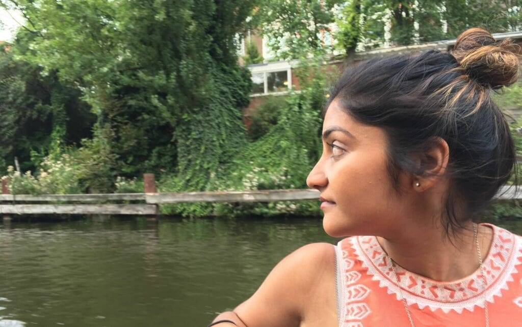 Aditi, a young person with dark hair and blonde highlights in their bun, sits in front of water with green trees in the background. They are turned in profile and are wearing an orange tank top with white patterned embroidery.