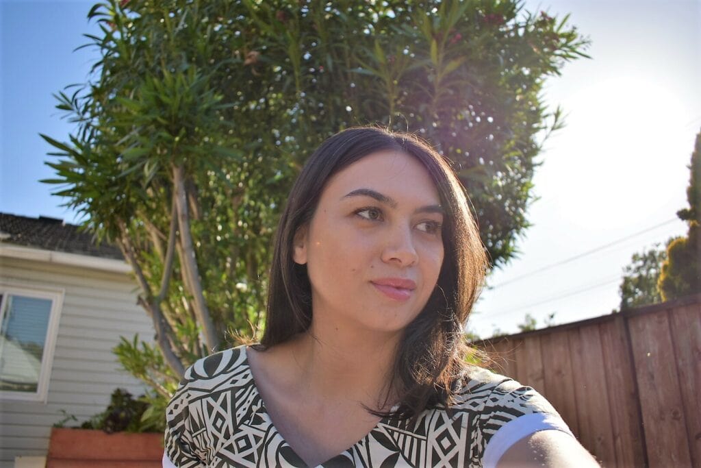 Betsy, a woman with brown hair and tan skin, is shown in front of a tree and blue sky