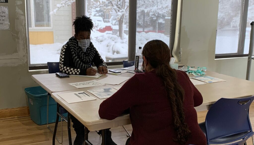 Two people sit across a table from one another. The table has lots of paperwork on it. Outside past the windows, snow covers the ground.