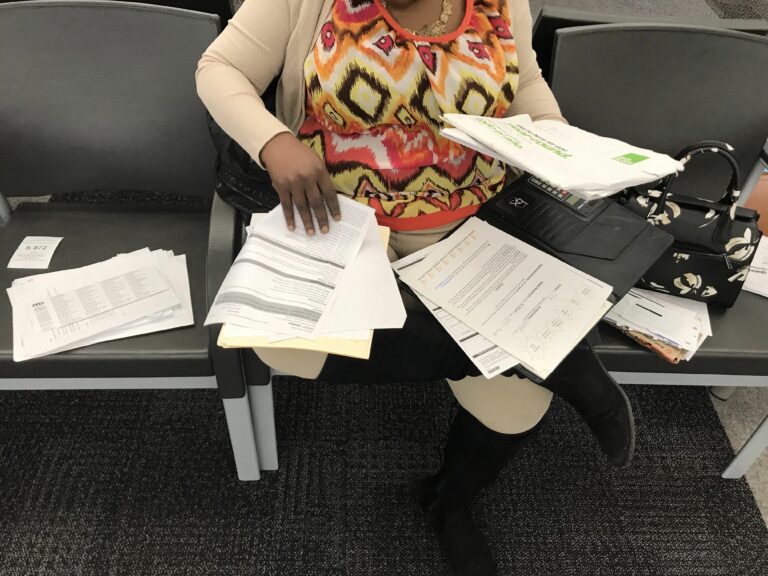 A woman in a square patterned shirt sits in an administrative office with dozens of documents spread across her lap and the chairs next to her