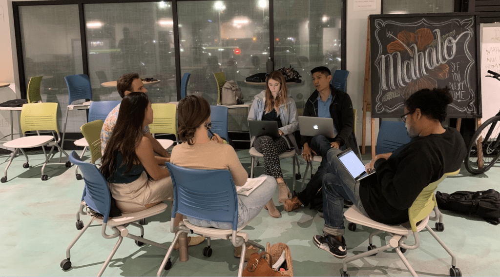 A group of people sit around in a circle with laptops open in their laps. Behind them is a chalkboard that says