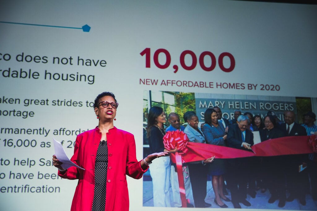 Maria Benjamin stands on stage at the Code for America summit in 2018 wearing a red blazer