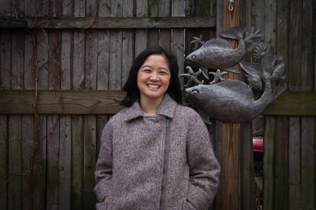 Mari Nakano stands in front of a wooden fence with an ornamental metal decal showing two fish and several starfish. Mari is smiling, has shoulder length black hair, and is wearing a grey coat.