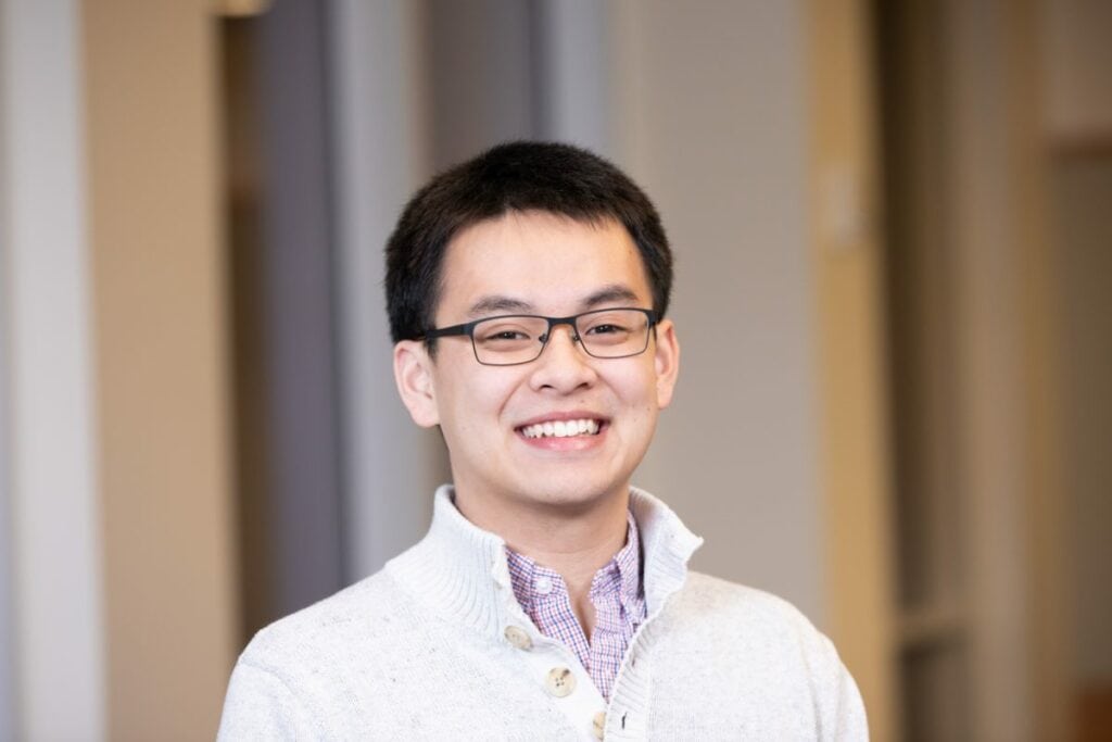 Chris Kuang is smiling in front of a soft background. He wears a white collared sweater and has black frame glasses.