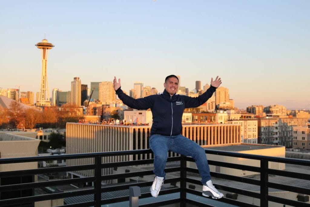 Victor sits on a rooftop ledge with his arms outstretched. He is smiling and wearing a Code for America jacket. Behind him to his right is the Seattle space needle