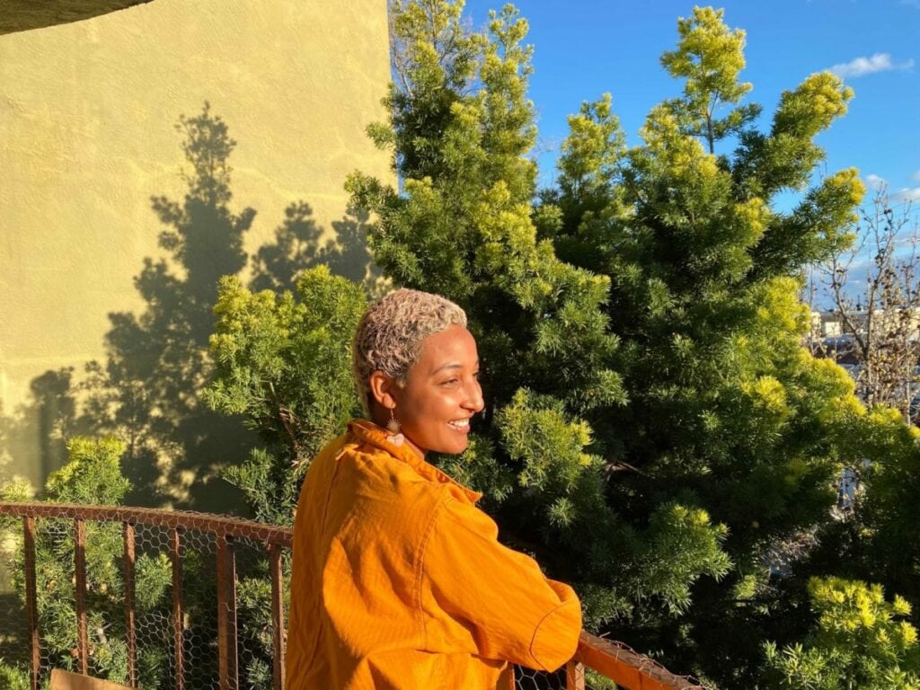 Symonne stands in front of a tree full of green leaves. They have short blonde hair and are wearing an orange shirt