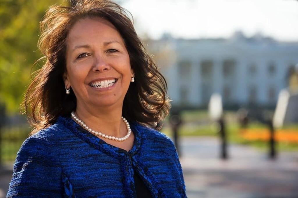 Victoria Vasquez smiles in front of a white building. She wears a pear necklace and a blue sweater