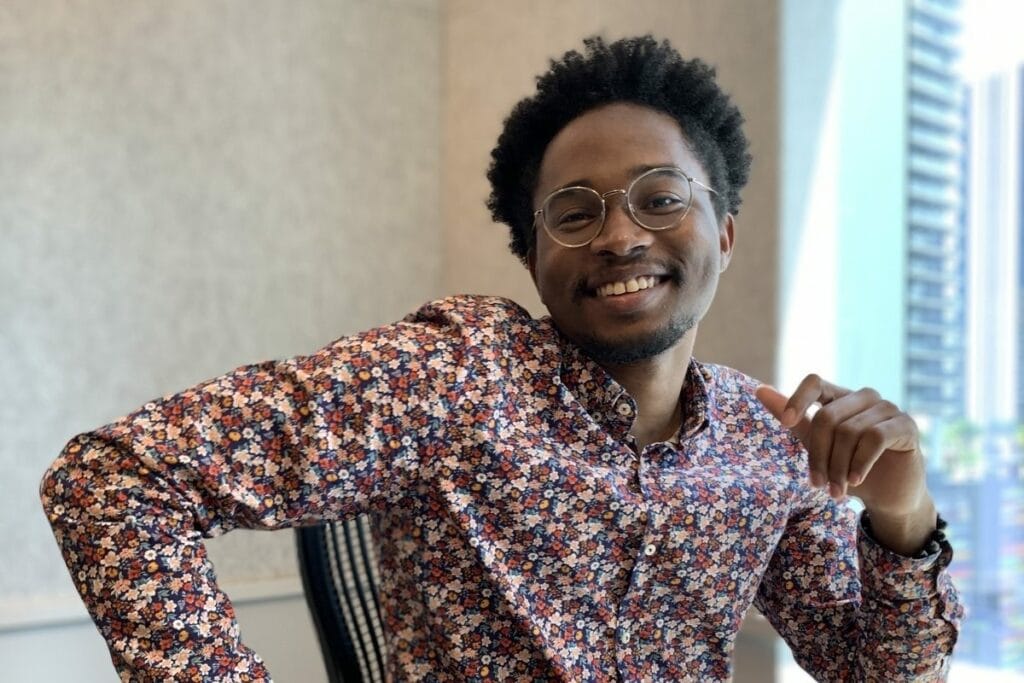 Gregory Johnson sits in a chair with his right elbow up. He is wearing a floral patterned shirt and wire framed glasses