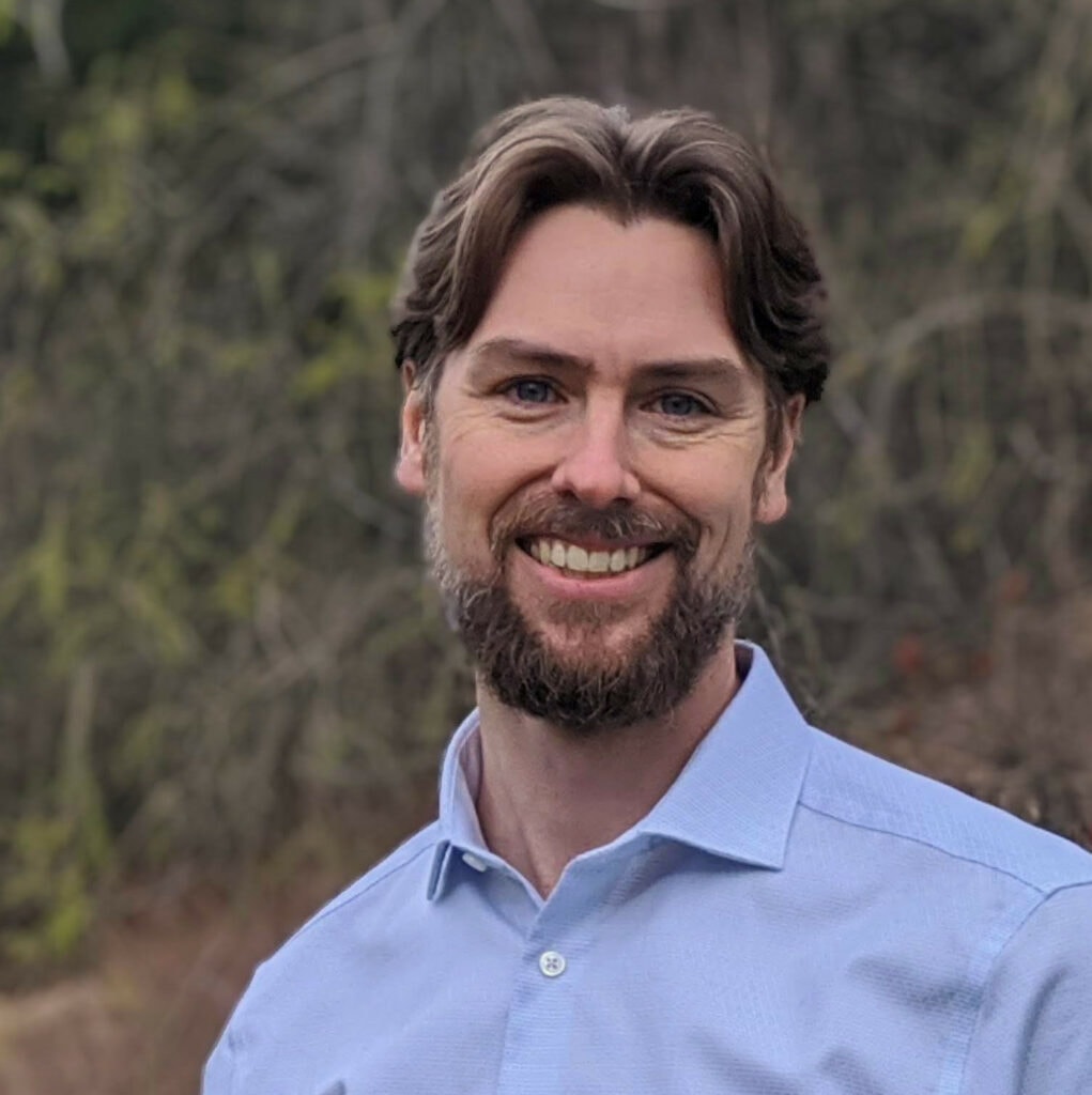 Micah stands against a backdrop of greenery wearing a blue collared shirt