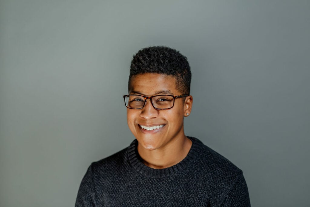 Simone stands against a grey background, wearing black glasses and a black sweater.