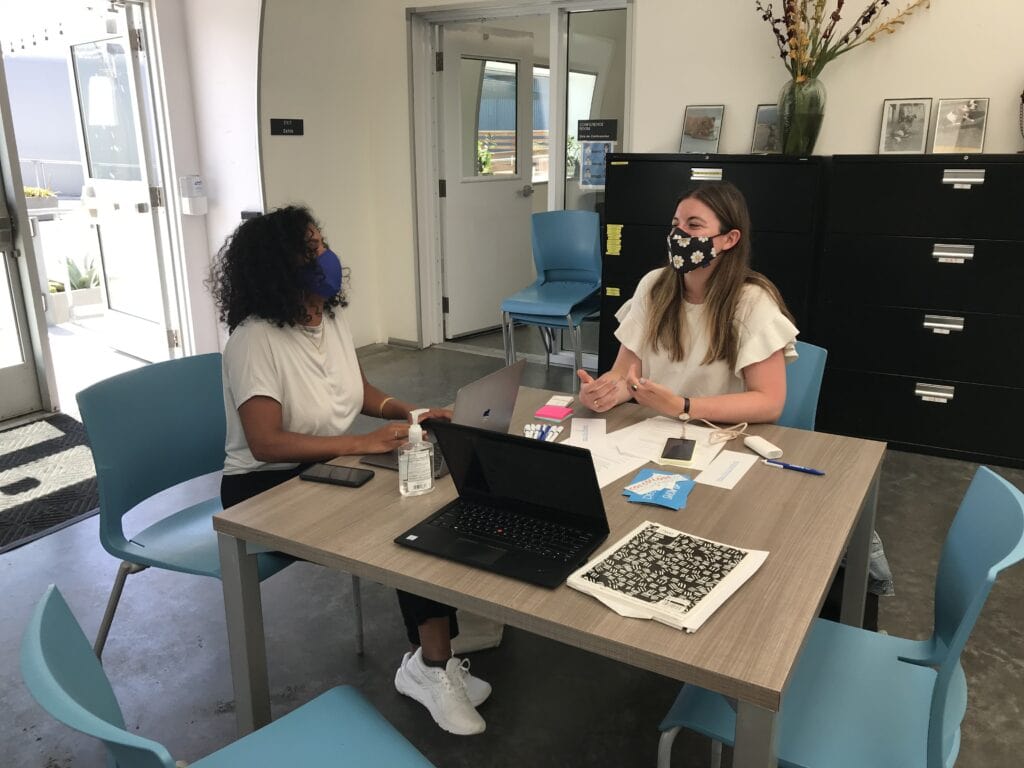 Two people wearing masks sit at a table with. their computers and post it notes