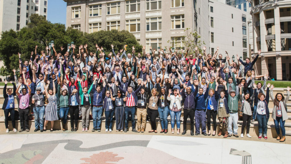 Group photo of Brigade members attending Summit 2019. They wave their hands in the air with trees and buildings in the background.